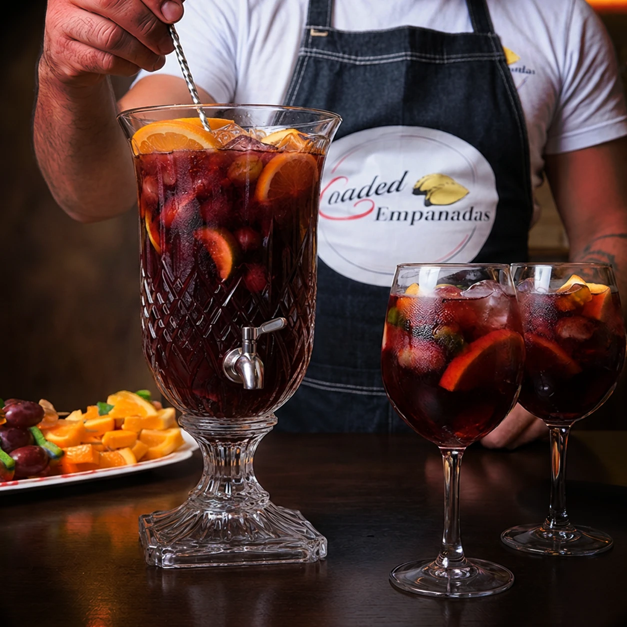 A bartender in a Loaded Empanadas apron stirring a crystal dispenser of red sangria beside two fruit-filled glasses